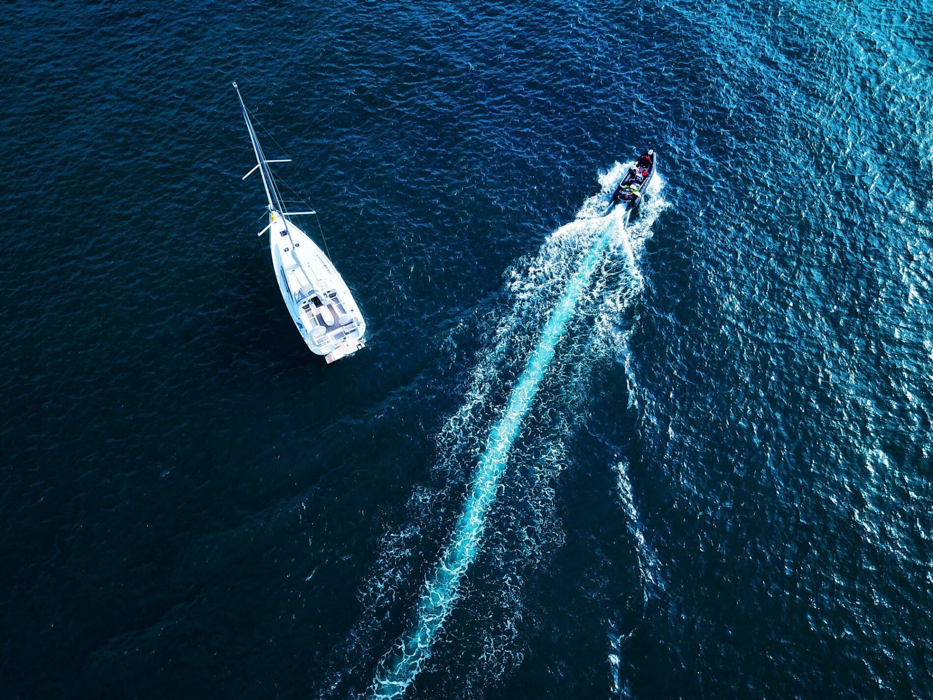 The image depicts a serene marina filled with various types of boats and super yachts. The water in the marina is calm, reflecting the clear, blue sky above. On the left side of the image, there is a cluster of smaller boats, which appear to be sailboats and pleasure crafts. Each of these boats is moored to wooden docks that stretch out into the water.

Towards the centre of the image, the size of the boats increases, transitioning to larger sailing yachts and motor yachts. These vessels have sleek, modern designs with white hulls that glisten under the sunlight.

To the right side of the image, the yachts reach an impressive scale, with several super yachts docked prominently. These luxurious vessels are equipped with multiple decks, expansive windows, and intricate details, showcasing their grandeur and opulence.

In the background, there's a picturesque landscape featuring lush green trees and hills, adding a natural frame to the marina. The overall atmosphere of the image is tranquil and reflective, capturing the elegance and lifestyle associated with maritime leisure and luxury.