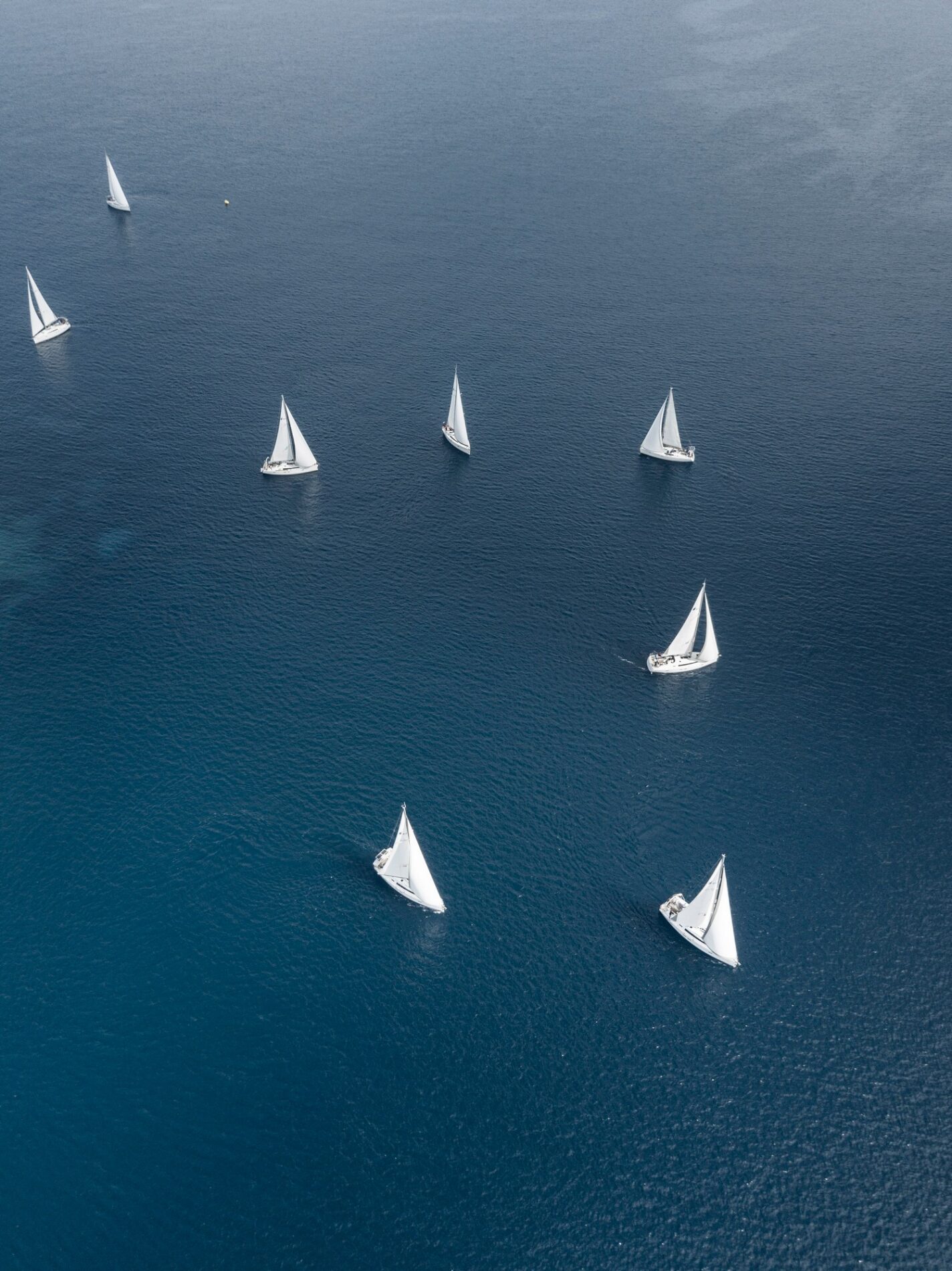 The image displays a close-up scene of several small, colorful paper boats floating on the surface of a gently rippling body of water. The boats are made of folded paper in various bright, vivid colours, including red, blue, yellow, and green. The reflection of the boats on the water adds a picturesque quality to the composition. The water around the boats is clear, with slight undulations creating a serene and peaceful ambiance. In the background, out of focus, are hints of greenery that suggest the scene might be set in an outdoor environment, possibly near a garden or park. The overall mood of the image is calm and nostalgic, evoking memories of childhood play and simple, creative outdoor activities.